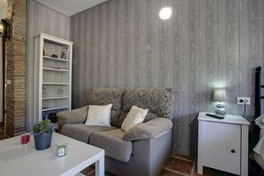 Um quarto aconchegante com um sofá cinzento, uma mesinha de cabeceira branca com abajur, uma estante de livros e um vaso com planta.