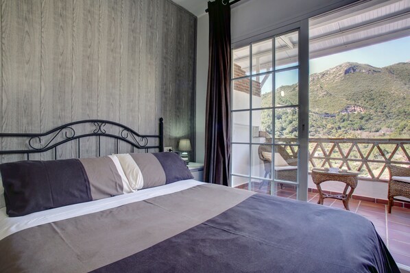 Quarto com cama grande, vista para montanhas por uma varanda e uma cadeira de vime.