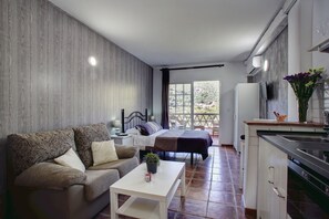 Quarto moderno com sofá, cama, mesinha com planta, área de cozinha e janela grande com vista para o verde.
