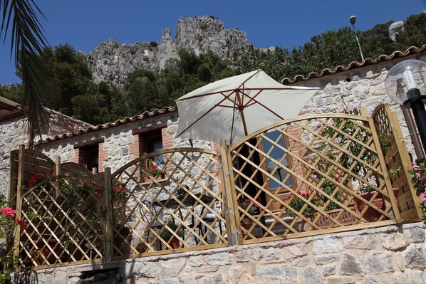 A stone building with a rustic charm, featuring a wooden gate with a lattice design, a white umbrella providing shade, and a view of a rocky hill in the background.