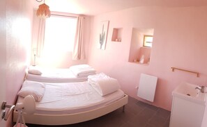 A bedroom with two beds, a window with curtains, a bedside lamp, a mirror, a radiator, and a sink.