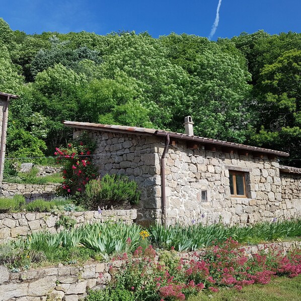 A stone building with a tiled roof, surrounded by lush greenery and flowering plants.