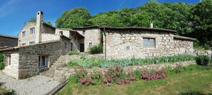 A stone building with multiple windows and a chimney, surrounded by a garden with various plants and flowers.