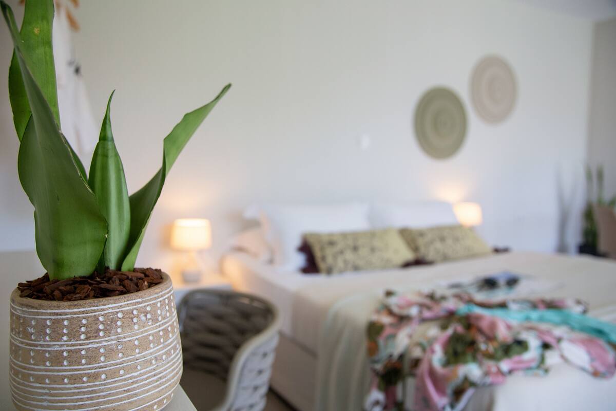 Un dormitorio con cama, almohadas, una mesita de noche y una planta en maceta.