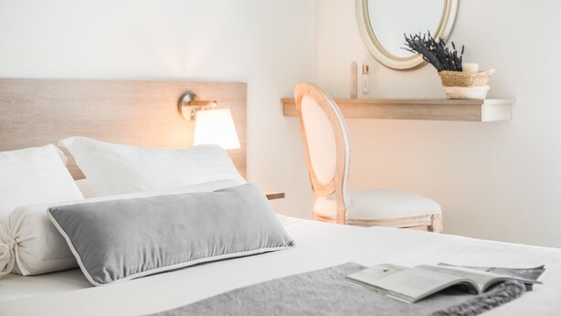 Una cama bien hecha con almohadas, una lámpara de noche, una silla, una estantería con un espejo y un libro.