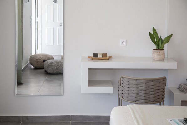 Ein modernes Zimmer mit weißer Wand, einem weißen Regal mit einem Topf und einer Pflanze, einem gewebten Sessel und einem Bett mit weißer Bettdecke.