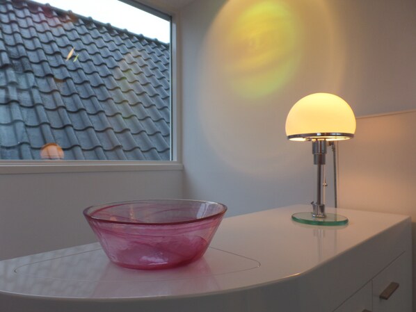A pink glass bowl on a white surface next to a lamp with a yellow shade, with a tiled roof visible through a window.