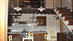 A kitchen with wooden cabinets, a dining table, and chairs. There is a staircase with a railing leading to an upper level. - Apartment for 6 with 3 rooms near San Gimignano and Volterra. (montaione)