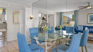 Een moderne eethoek met een glazen tafel en blauwe fauteuils, een plafondventilator en uitzicht op de woonkamer.