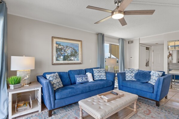 A living room with a blue sofa set, a ceiling fan, a framed painting, and a side table with a lamp. - Cajun Fun 1060 - 1 Br Condo (Destin)