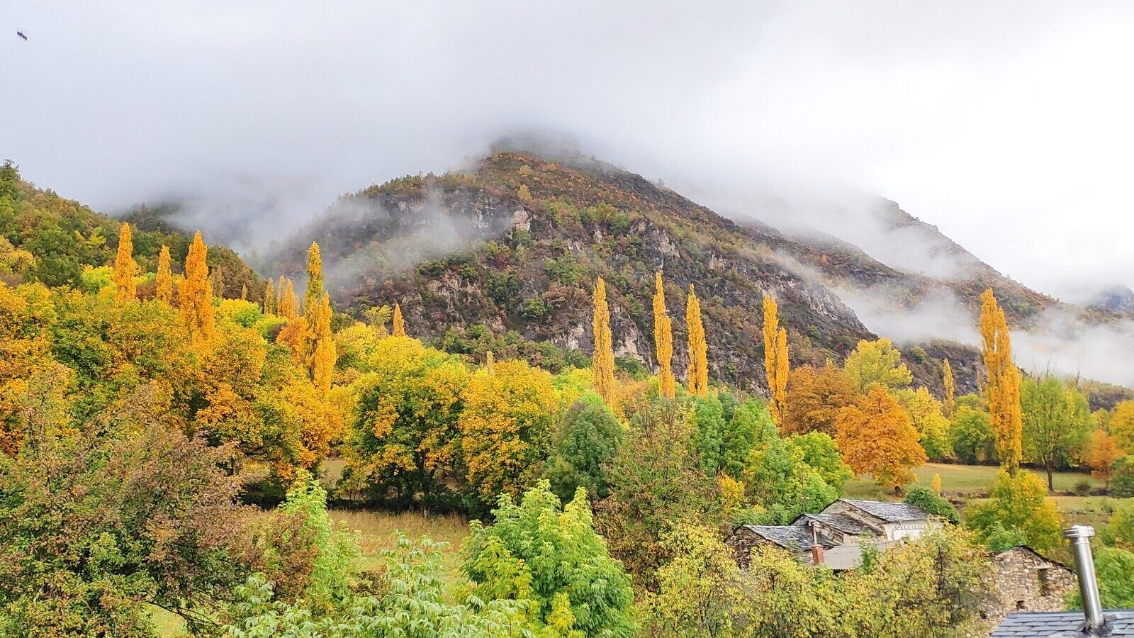 Paisaje montañoso con follaje otoñal y un pequeño edificio en primer plano.
