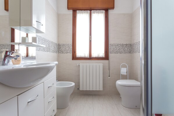 Un baño moderno con un lavamanos, inodoro y bañera blancos. Hay una ventana con cortinas y un radiador sobre el inodoro.
