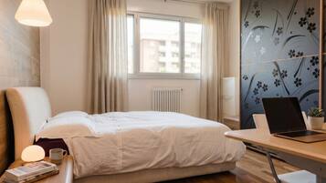 A hotel room with a bed, a desk with a laptop, a window with curtains, and a radiator.