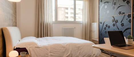 A hotel room with a bed, a desk with a laptop, a window with curtains, and a radiator.