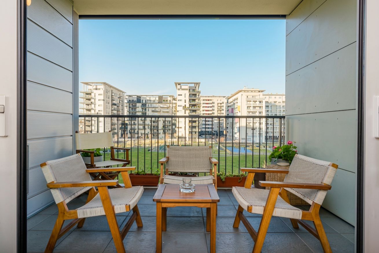 A balcony with wicker chairs and a table, overlooking a residential area with buildings.