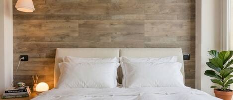 A bedroom with a wooden headboard, white bedding, a bedside lamp, and a potted plant.