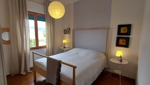 Une chambre à coucher avec un lit, une fenêtre avec des rideaux, une table de chevet avec une lampe et un cadre photo fixé au mur.