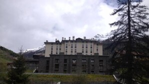 Un immeuble à plusieurs étages, doté d’une façade symétrique, qui ressemble probablement à un hôtel ou à un complexe touristique, se dressant sur un fond de montagnes et d  ’un ciel nuageux.