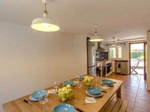 A dining area with a long wooden table set for four, blue plates, and a bowl of apples. There are hanging pendant lights above.