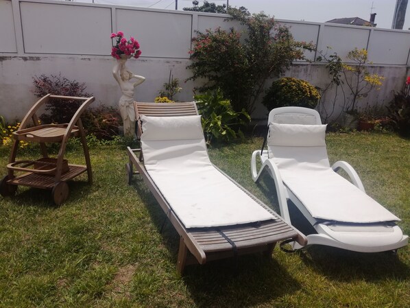 Dos tumbonas con cojines blancos, un carrito de madera y una estatua adornada con flores rosadas.