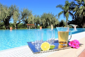 Escena junto a una piscina con agua azul cristalina, una bandeja con vasos, una jofaina de líquido amarillo, una rodaja de lima y flores rosadas.