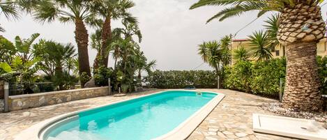A rectangular swimming pool with a stone tile deck, surrounded by palm trees and a stone wall.