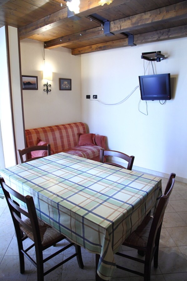 A dining area with a table covered by a plaid tablecloth, surrounded by wooden chairs. A red plaid sofa is against the wall, and a television is mounted above it.