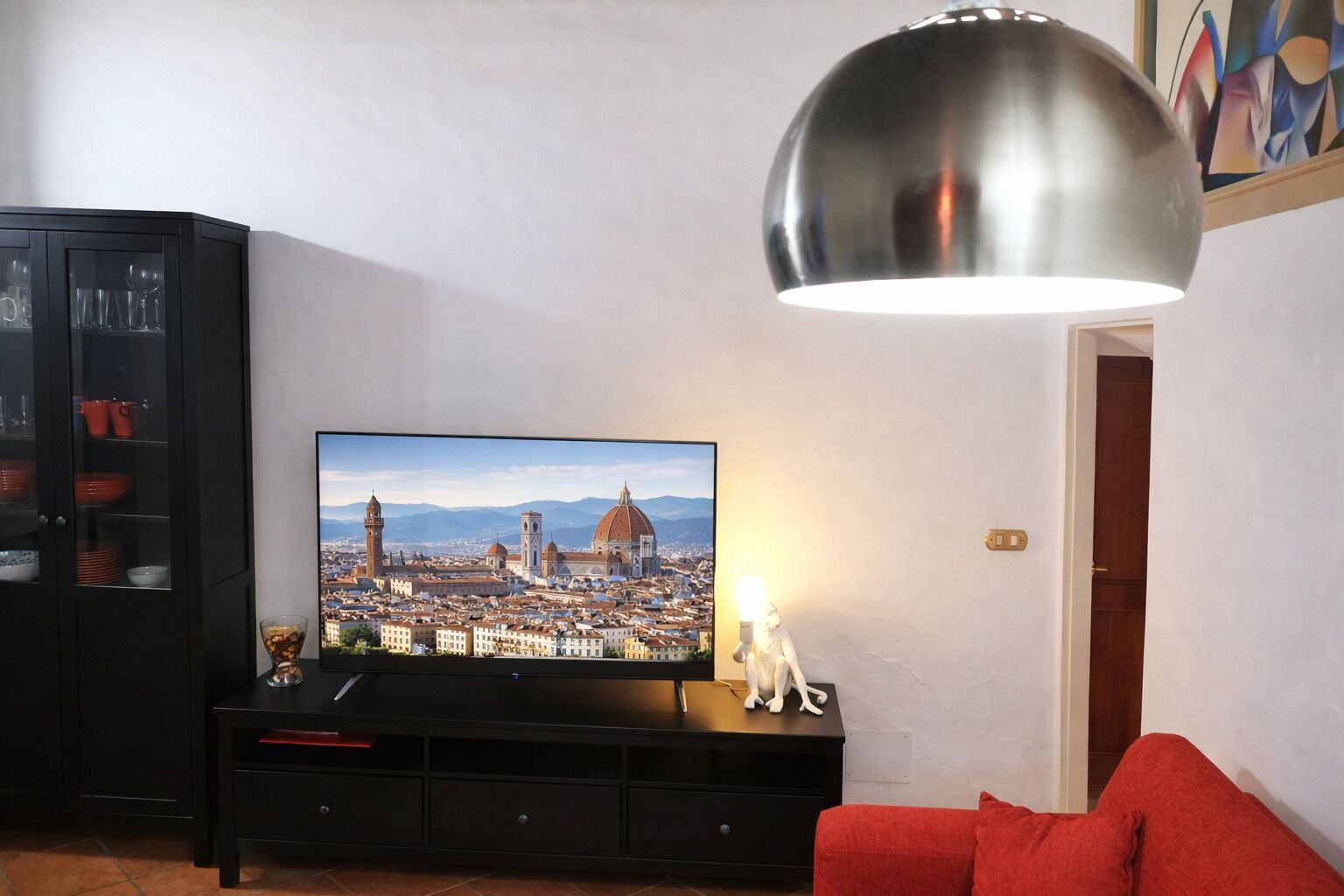 A living room with a flat-screen TV displaying a cityscape, a black entertainment unit, a red sofa, and a large pendant light.