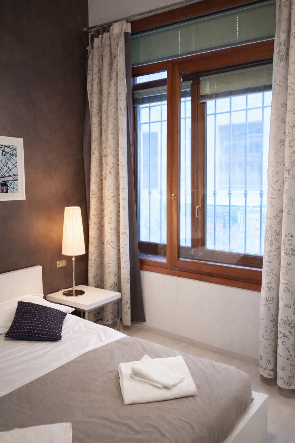 A hotel room with a bed, a bedside table with a lamp, a window with curtains, and a view of the outside.