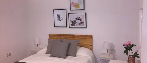 A bedroom with a wooden headboard, a bed with white bedding and a dark throw, two bedside lamps, a nightstand with a vase of flowers, and wall-mounted artwork.