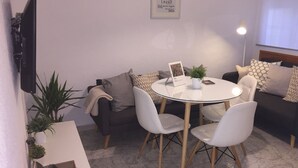 Un saló d'estar acollidor amb una taula rodona de vidre envoltat de cadires blanques, un sofà fosc amb coixinets estampats, una televisió plana penjada a la paret i una citació inspiradora emmarcada.