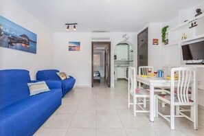 A modern living room with a blue sofa, a dining area with white chairs, and a kitchen in the background.