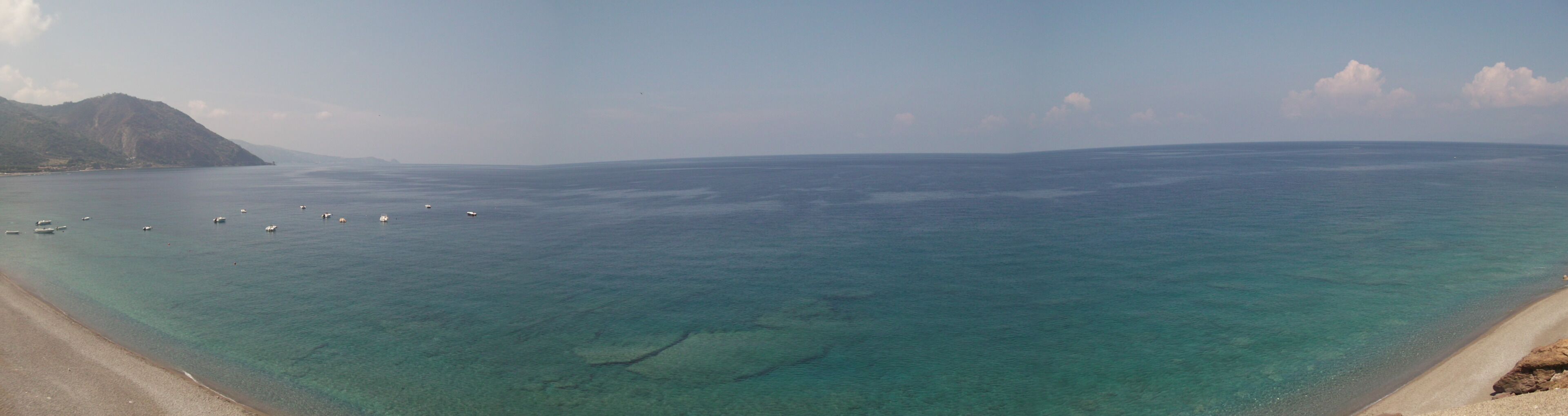 A coastal beach with clear turquoise waters, boats, and a mountainous backdrop.