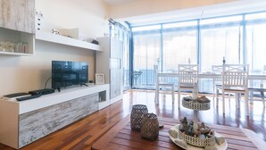 A modern living room with a wooden floor, a white entertainment unit with a flat-screen TV, and a dining area with white chairs and a table.