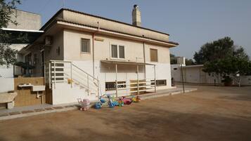 Two-story building with a flat roof and chimney, featuring a ground-floor balcony and a paved courtyard.