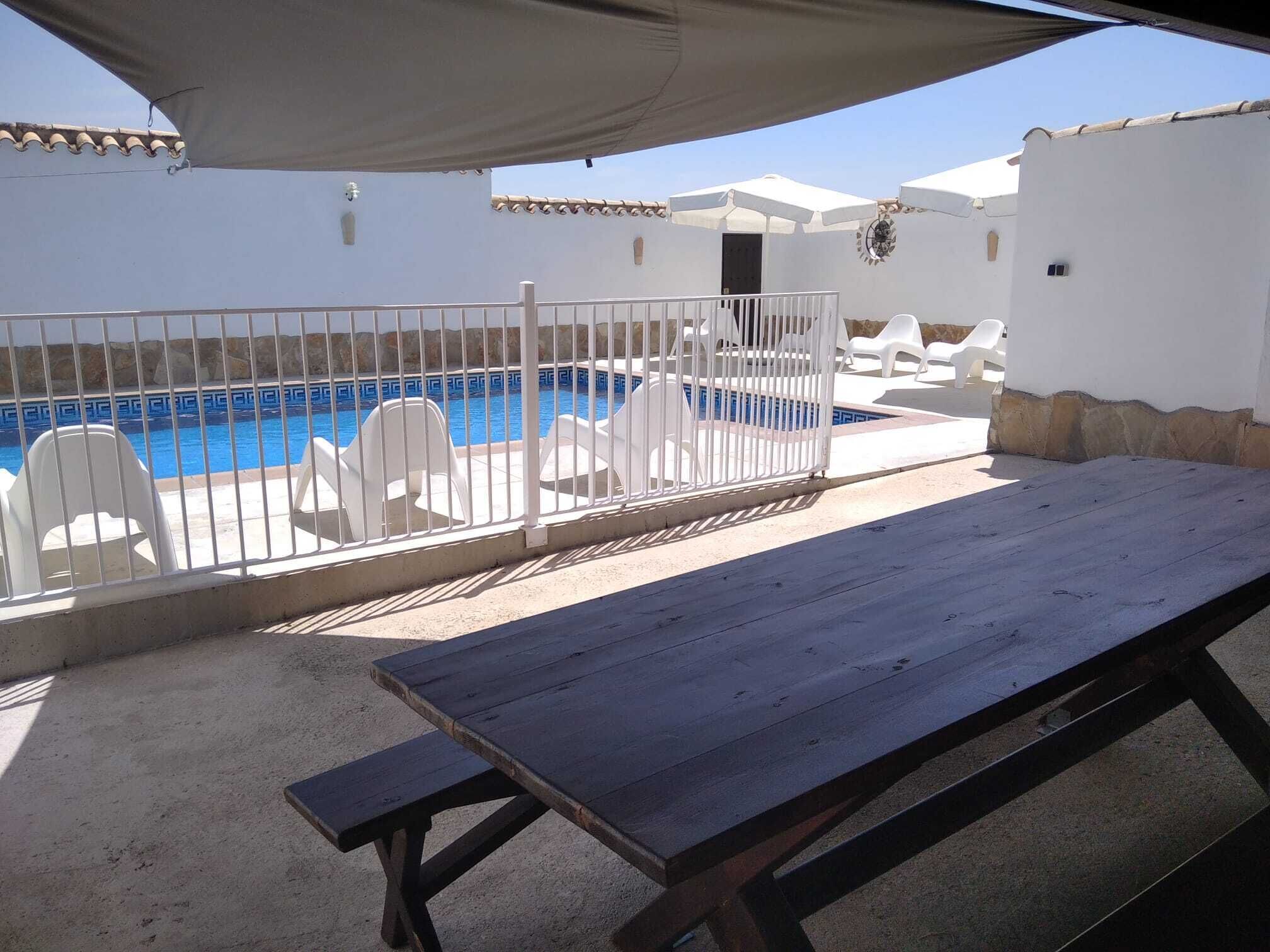 A poolside area with a wooden table and chairs, a white fence, and a covered seating area.