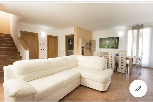 Uma sala de estar com sofá em formato de L, um espaço de jantar com mesa e cadeiras, uma escada e um quadro na parede.