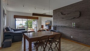 A modern living room with a wooden dining table, dark sofas, and a large window with a view. - Apartment with Garden and Jacuzzi. Sea view. Fully equipped. (Lagoa - Açores)