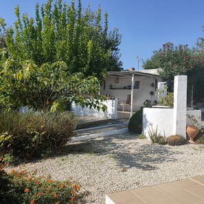 A white building with a covered porch, surrounded by greenery and a gravel path.