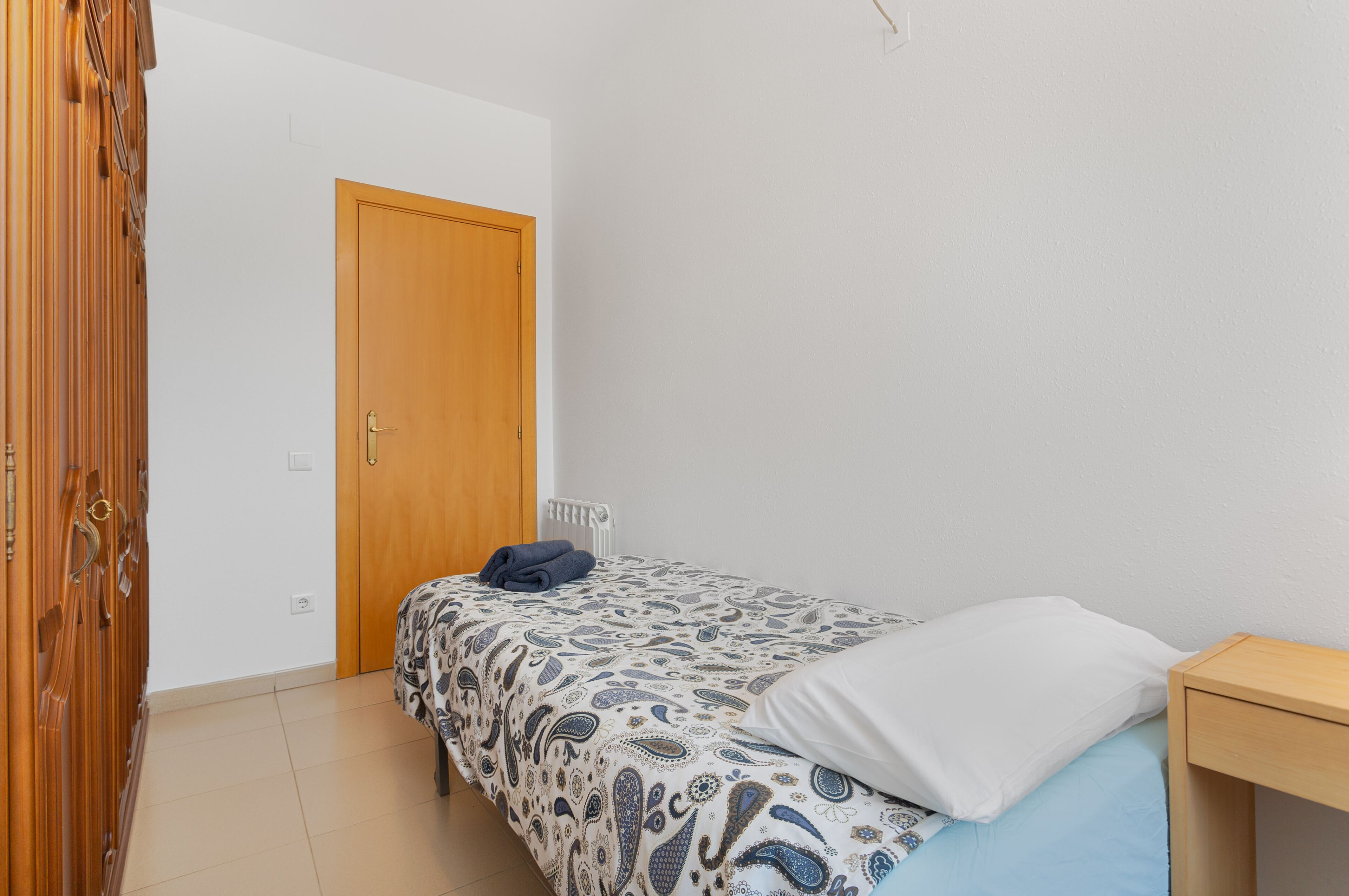 A bedroom with a bed, a wooden wardrobe, a white door, and a small bedside table.