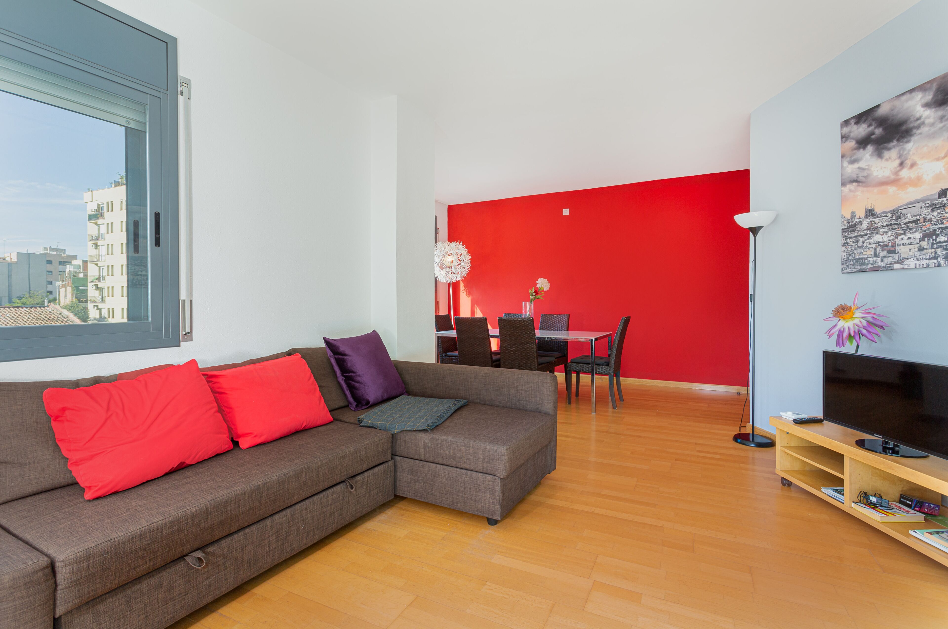 Un salon moderne avec un canapé d’angle gris, des coussins rouges, un coin repas avec un mur rouge et un téléviseur posé sur un meuble en bois.