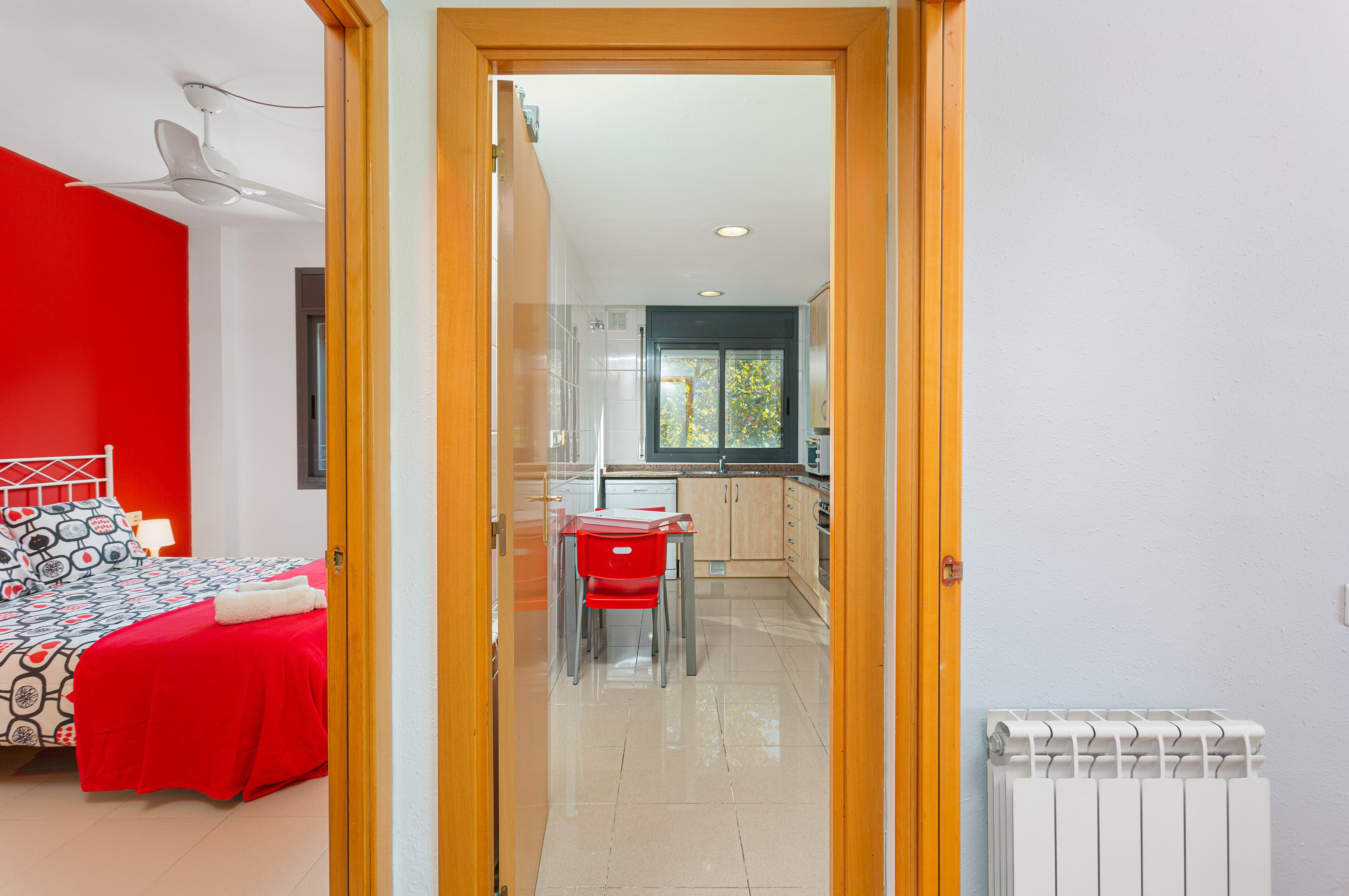 A room with a bed, a red chair, a small table, and a kitchen area with cabinets and a window.