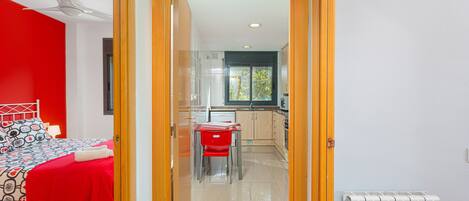 A room with a bed, a red chair, a small table, and a kitchen area with cabinets and a window.