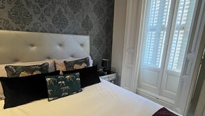 A bedroom with a tufted headboard, a large bed with white linens, multiple pillows, a dark red throw blanket, a nightstand with a lamp, and a window with blinds. - GRAN VIA MADRID (Madrid)