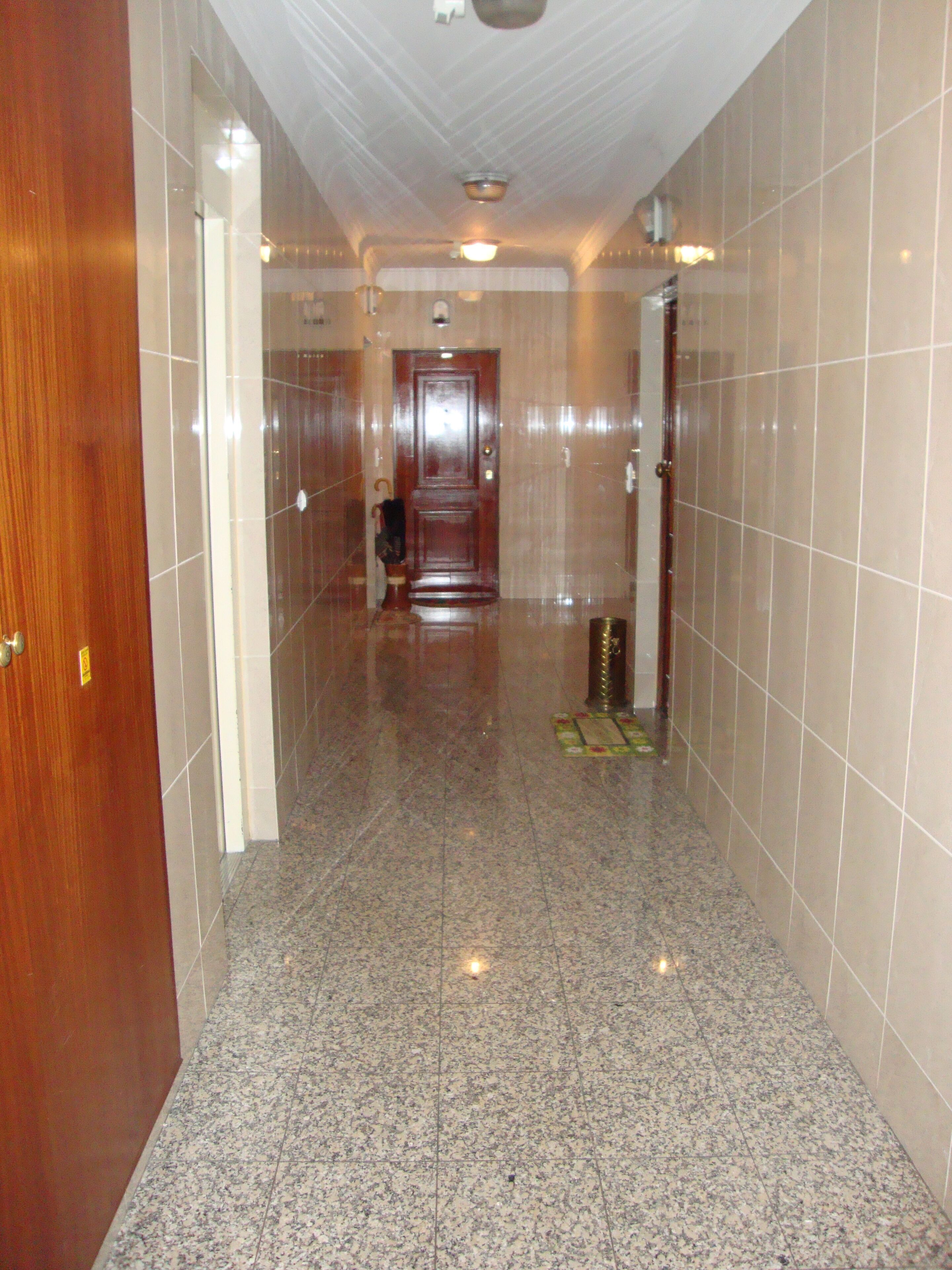 A hallway with a wooden door on the left, a tiled wall, and a tiled floor.