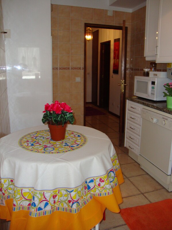 Une cuisine équipée d’un four à micro-ondes, d’un évier et d’un placard. On y trouve une table recouverte d’une nappe colorée et un pot de fleurs avec des fleurs rouges.