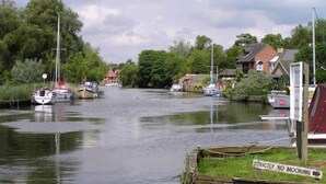 Un río tranquilo con barcos amarrados a la izquierda, un cartel que indica 'Strictly No Mooring' a la derecha y casas a lo largo de la orilla.