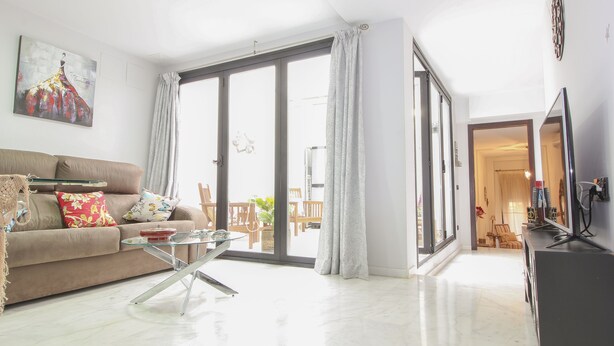 Un salón amplio con sofá, una mesa de centro de vidrio y un ventanal con cortinas.