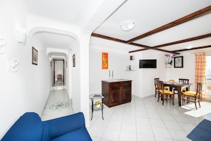 Un salon avec un canapé bleu, un coin repas avec des chaises en bois et une table, un téléviseur fixé au mur, et une petite table d’appoint avec un vase.