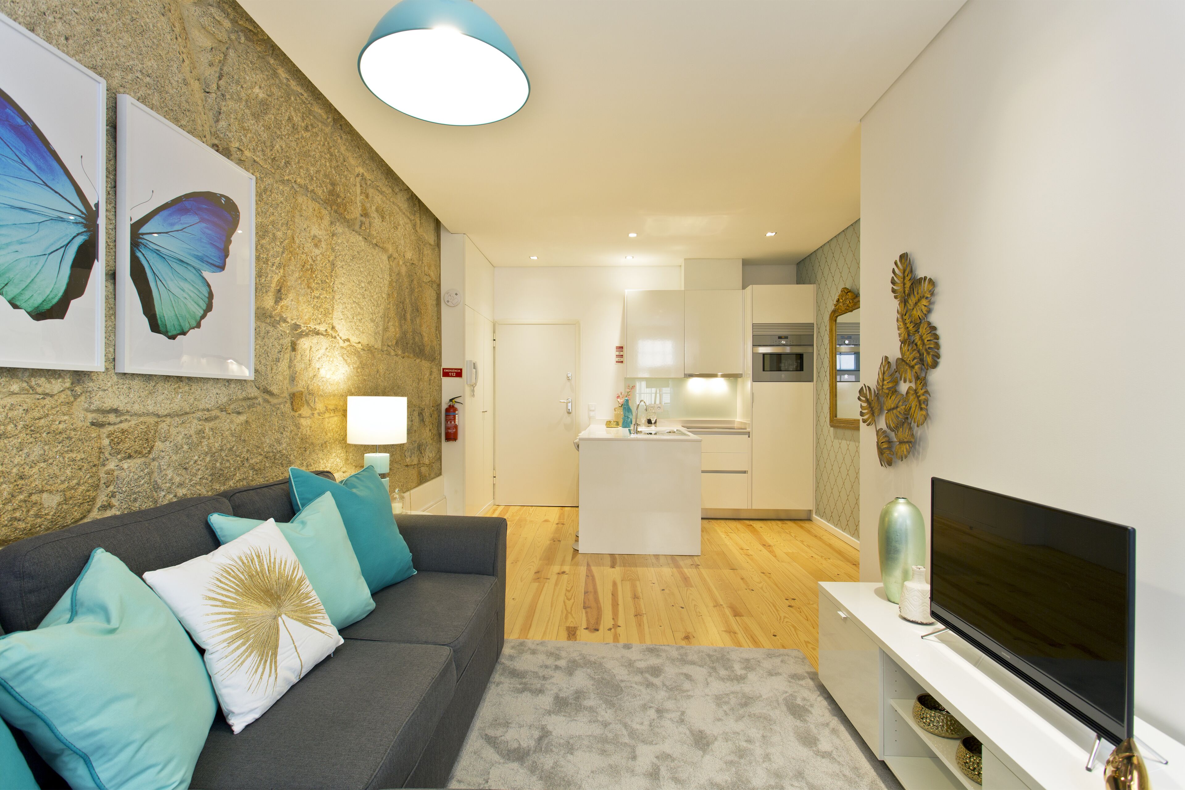 A modern living room with a grey sofa, teal cushions, a flat-screen TV, and a kitchen in the background.